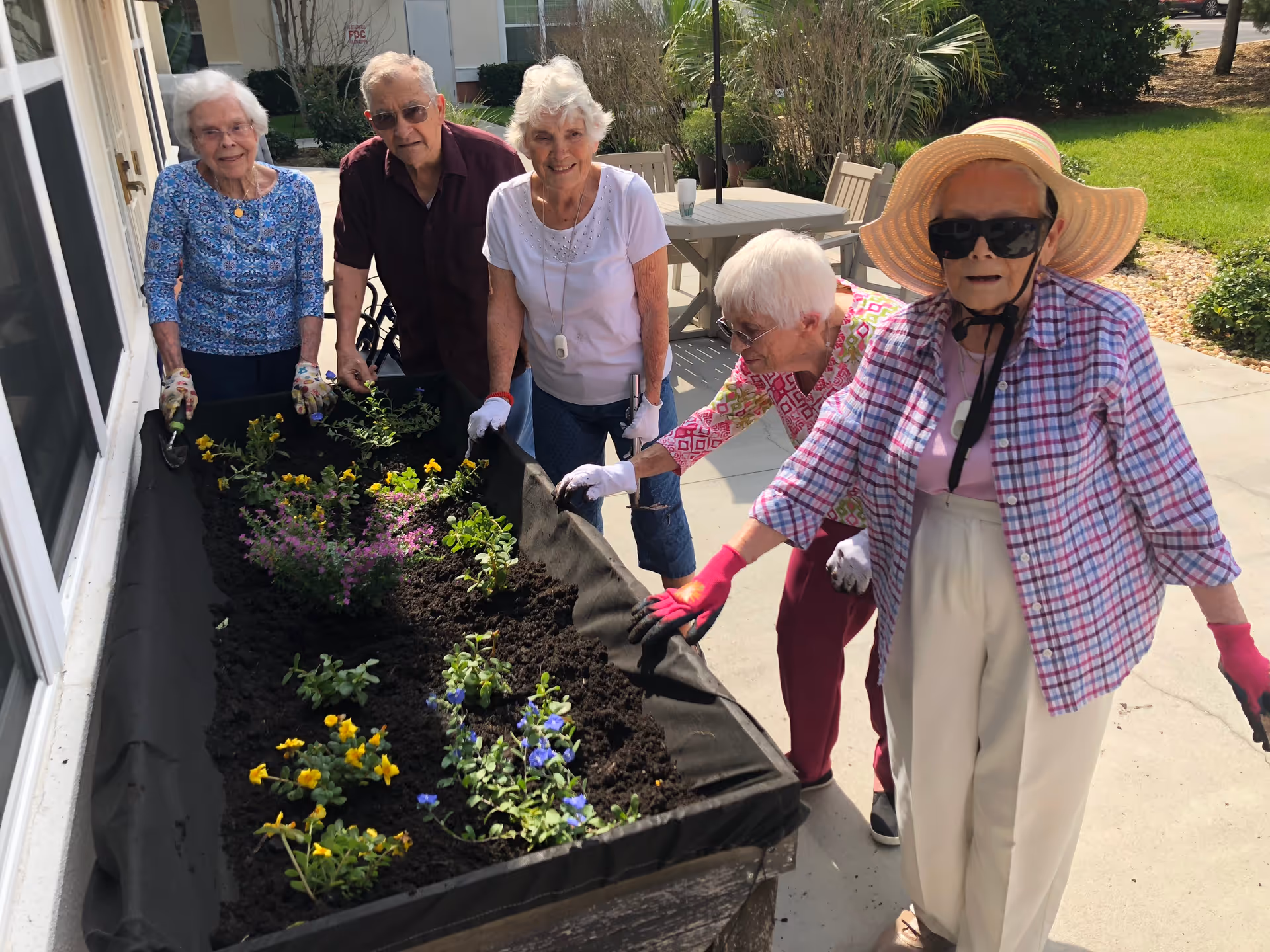 Five elderly individuals are gardening together outdoors beside a raised garden bed filled with soil and small flowering plants. They are smiling and wearing gardening gloves, with one person wearing a large sun hat and sunglasses. The setting appears to be a sunny day at a senior living facility.
