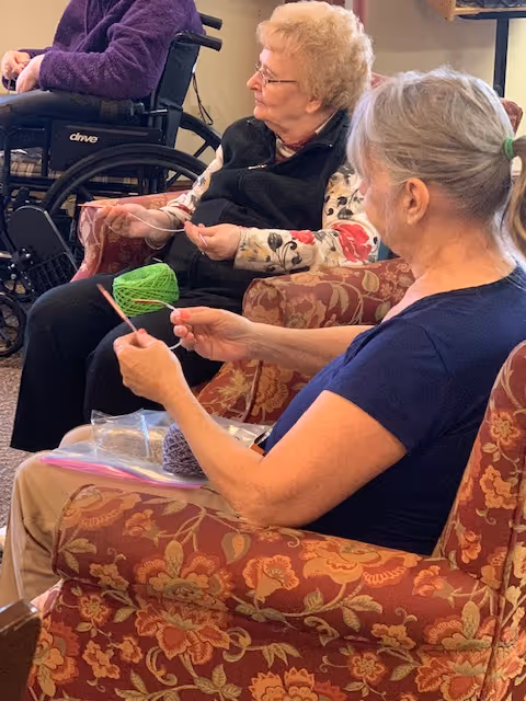 Two elderly women sitting in patterned armchairs in a common area, engaged in knitting with yarn and knitting needles. One woman has gray hair tied back and is wearing a navy blue shirt, while the other has short curly blonde hair and is wearing glasses and a black vest over a floral shirt. A person in a wheelchair is partially visible in the background.