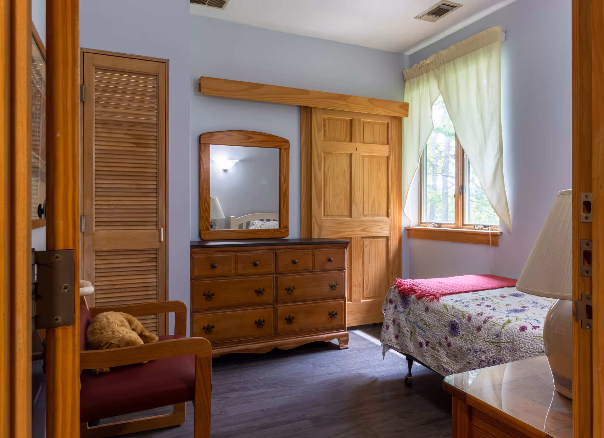 Cozy memory-care bedroom with wooden dresser, mirror, bed, chair, and window.