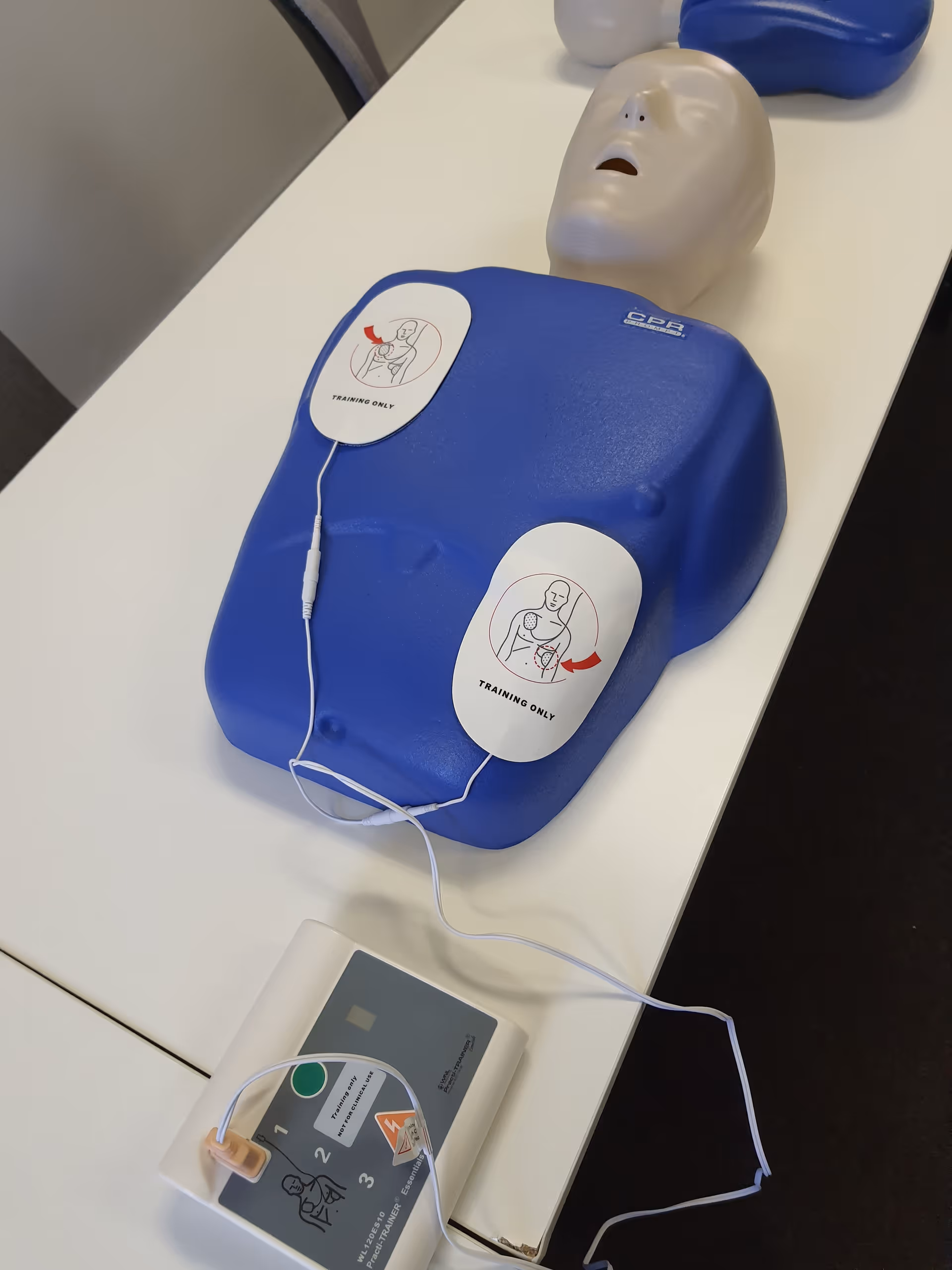 Blue CPR training mannequin with wired pads on a white table.