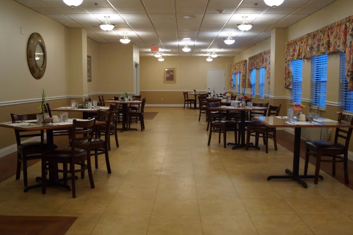 Interior view of a dining room with multiple wooden tables and chairs arranged neatly. Each table has place settings, glasses, and small flower arrangements. The room has beige walls with white trim, several windows with floral valances, and ceiling lights providing illumination.