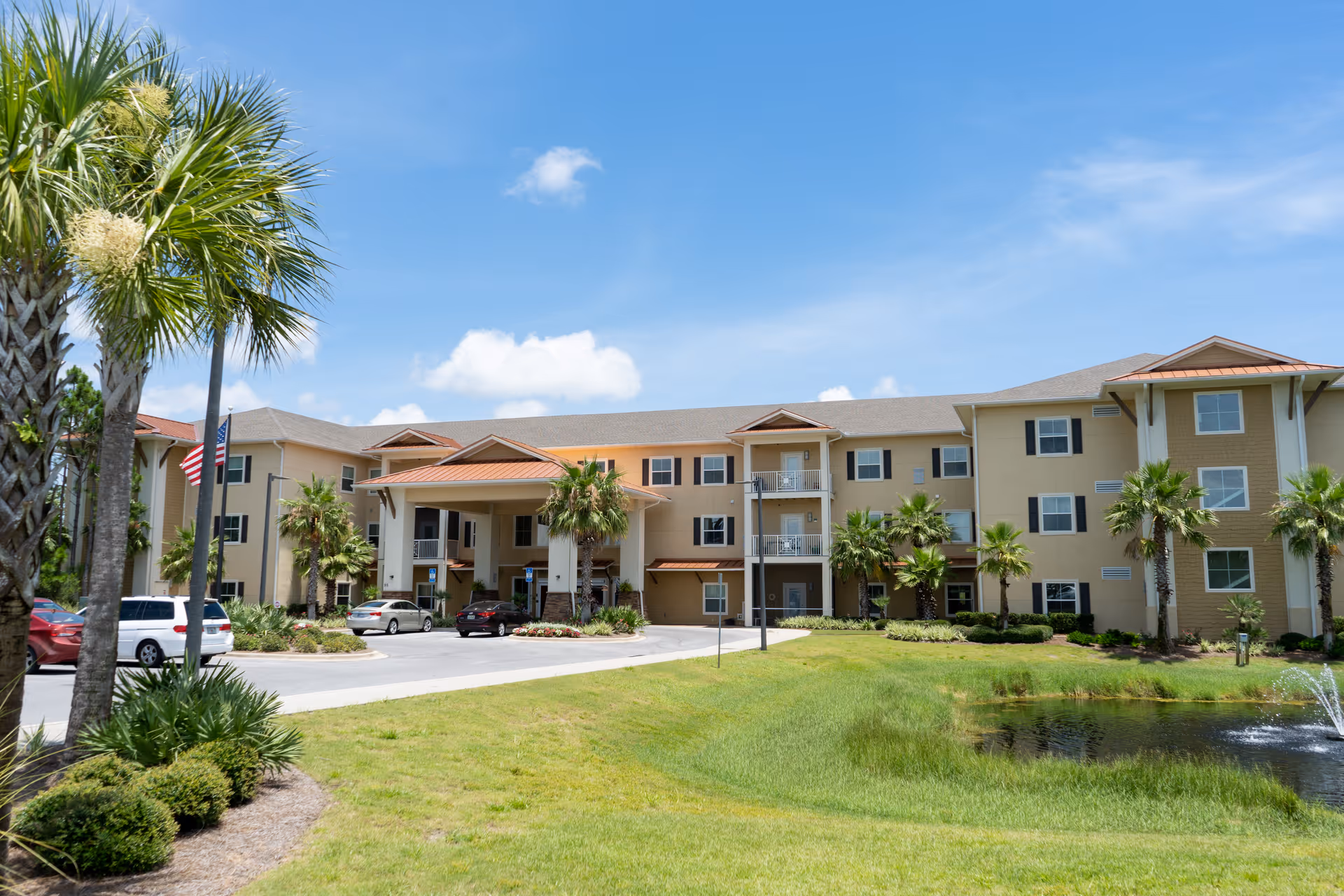 Exterior view of Superior Residences of Panama City Beach, a three-story senior living facility with a covered entrance, palm trees, a small pond with a fountain, and a clear blue sky.