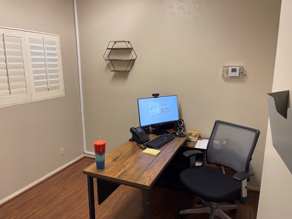 An office workspace with desk, computer, chair, tumbler, and wall shelf in a small room.