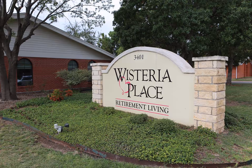 Exterior view of a Wisteria Place retirement living sign in landscaping near a brick building.