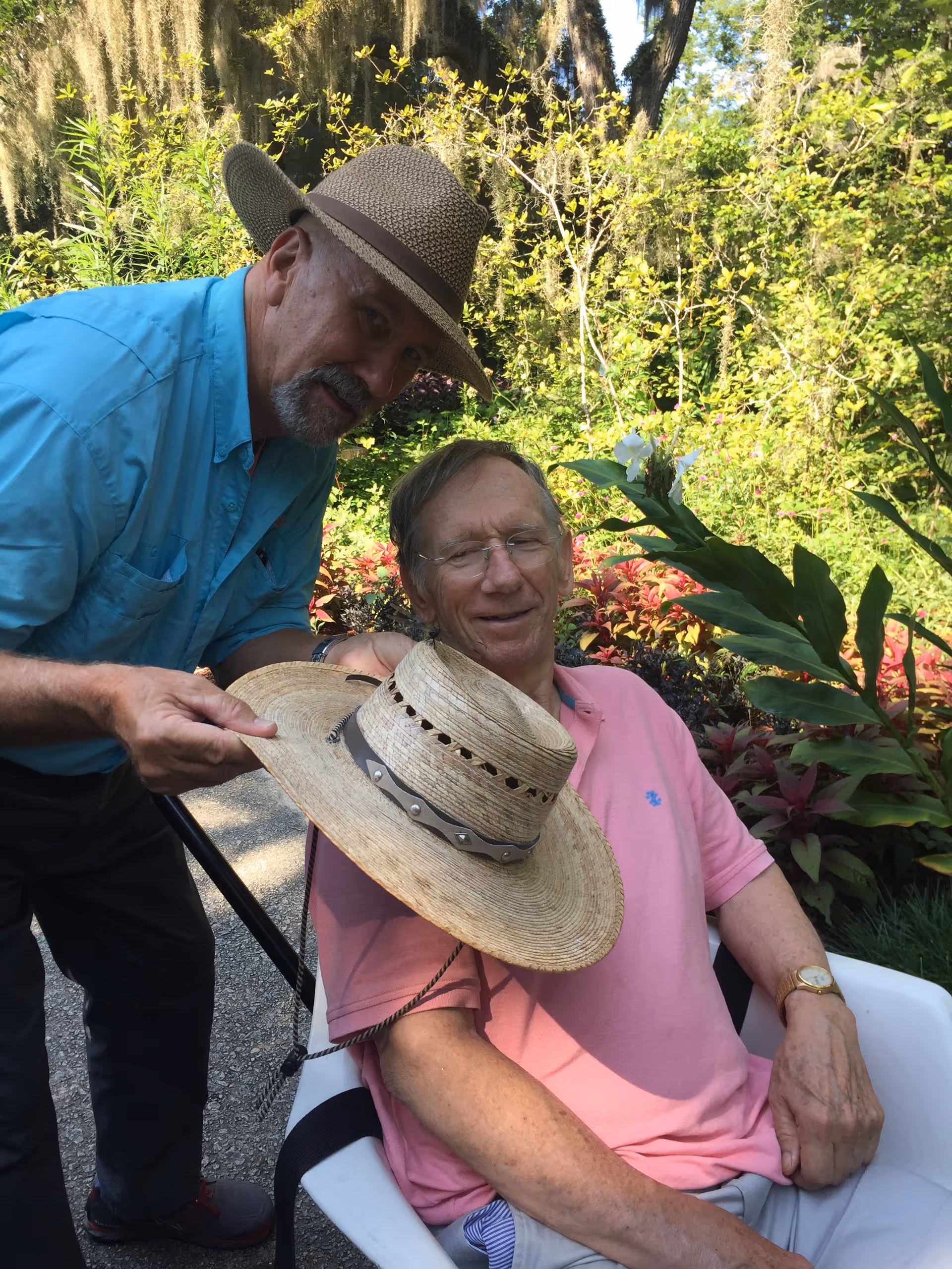 Two men outdoors in a garden; one sits in a chair, the other holds a hat.