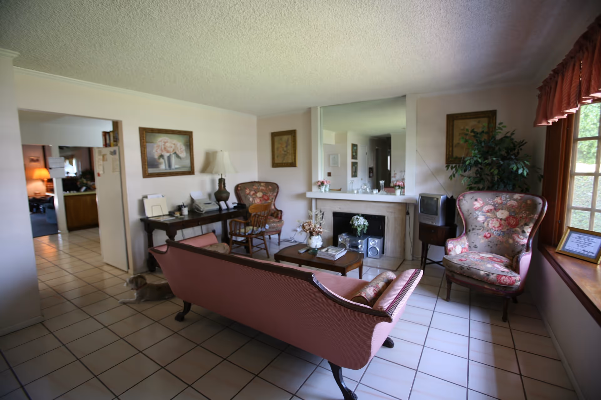 A cozy living room with a pink upholstered sofa, two floral armchairs, a wooden coffee table with books and a flower vase, a fireplace with decorative items on the mantel, a small TV on a stand, and a window with red curtains letting in natural light. There is a wooden desk with a lamp and papers, and a dog lying on the tiled floor near the entrance to an adjacent kitchen area.