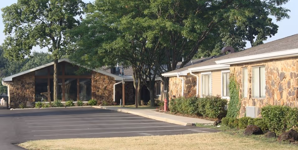 Exterior view of a single-story stone senior living facility with trees and parking.