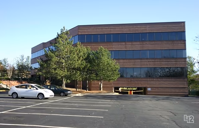 Three-story brick building with large windows, trees, and a parking lot.