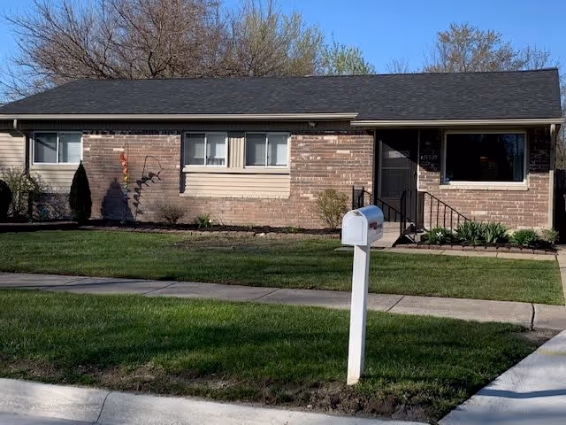 Single-story brick building with a dark shingled roof, several windows, a front door with steps and railing, a well-maintained lawn, a white mailbox on a post near the sidewalk, and trees in the background under a clear blue sky.