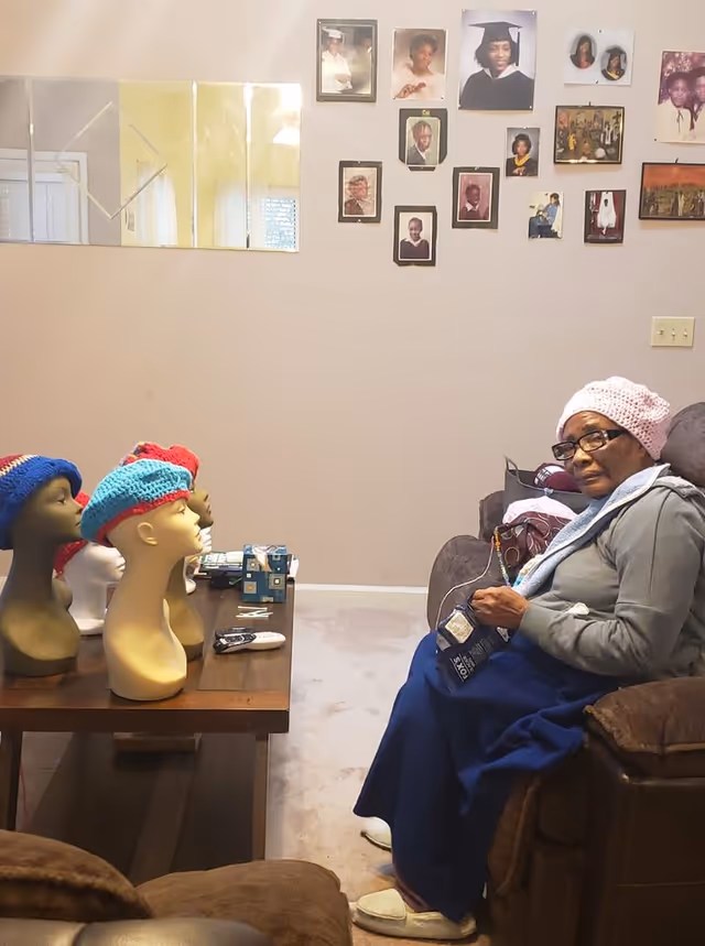 Resident sits in a living room chair beside a table with mannequin heads wearing hats.