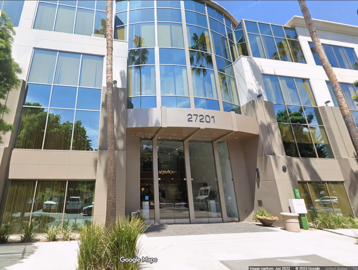 Modern glass-front building entrance with palm trees and a paved sidewalk.