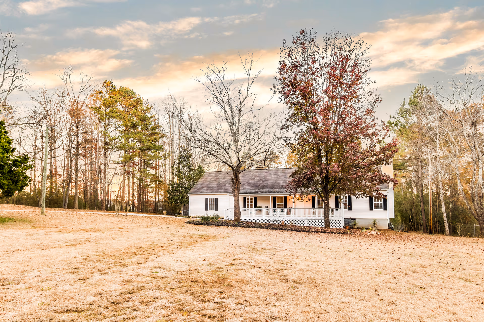 A white single-story house with a front porch surrounded by trees with autumn foliage, set on a large grassy area under a partly cloudy sky at sunset.