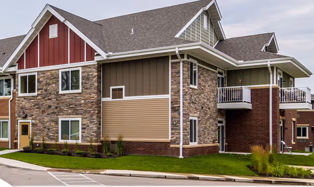 Exterior view of a two-story senior living facility building with a combination of stone, brick, and siding on the walls, multiple windows, a balcony, and a well-maintained lawn and landscaping.
