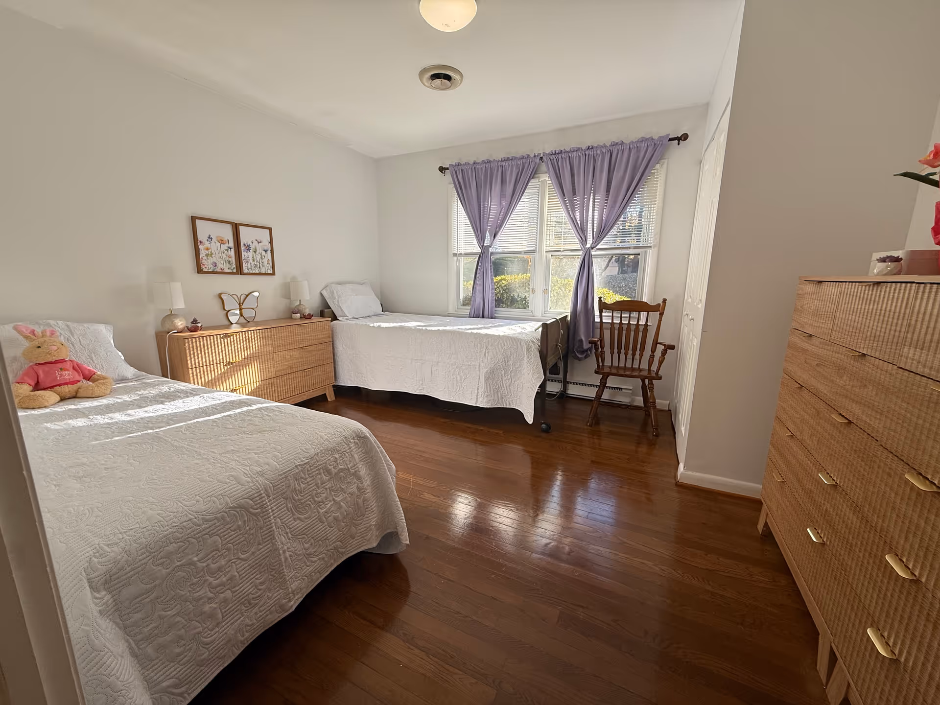 Two twin beds and dressers in a bright bedroom with purple curtains.
