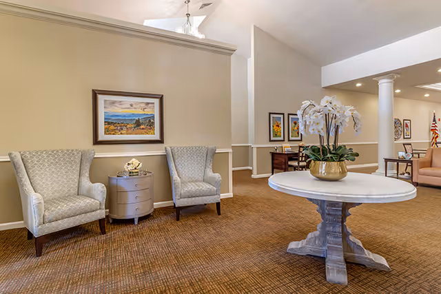 Spacious beige lobby with two patterned armchairs, a round pedestal table, and artwork.