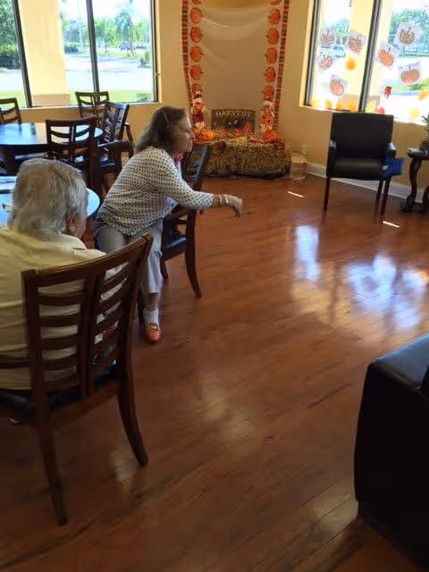 Two elderly women sit in a sunlit common room with wooden floors, chairs, tables, and fall decorations by the windows.