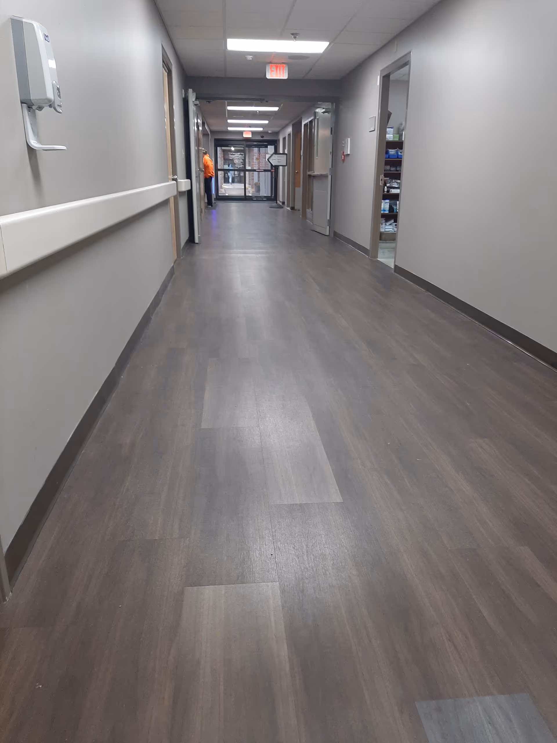 Long, empty hospital hallway with gray walls, wood flooring, handrails, and a distant orange-clad figure.