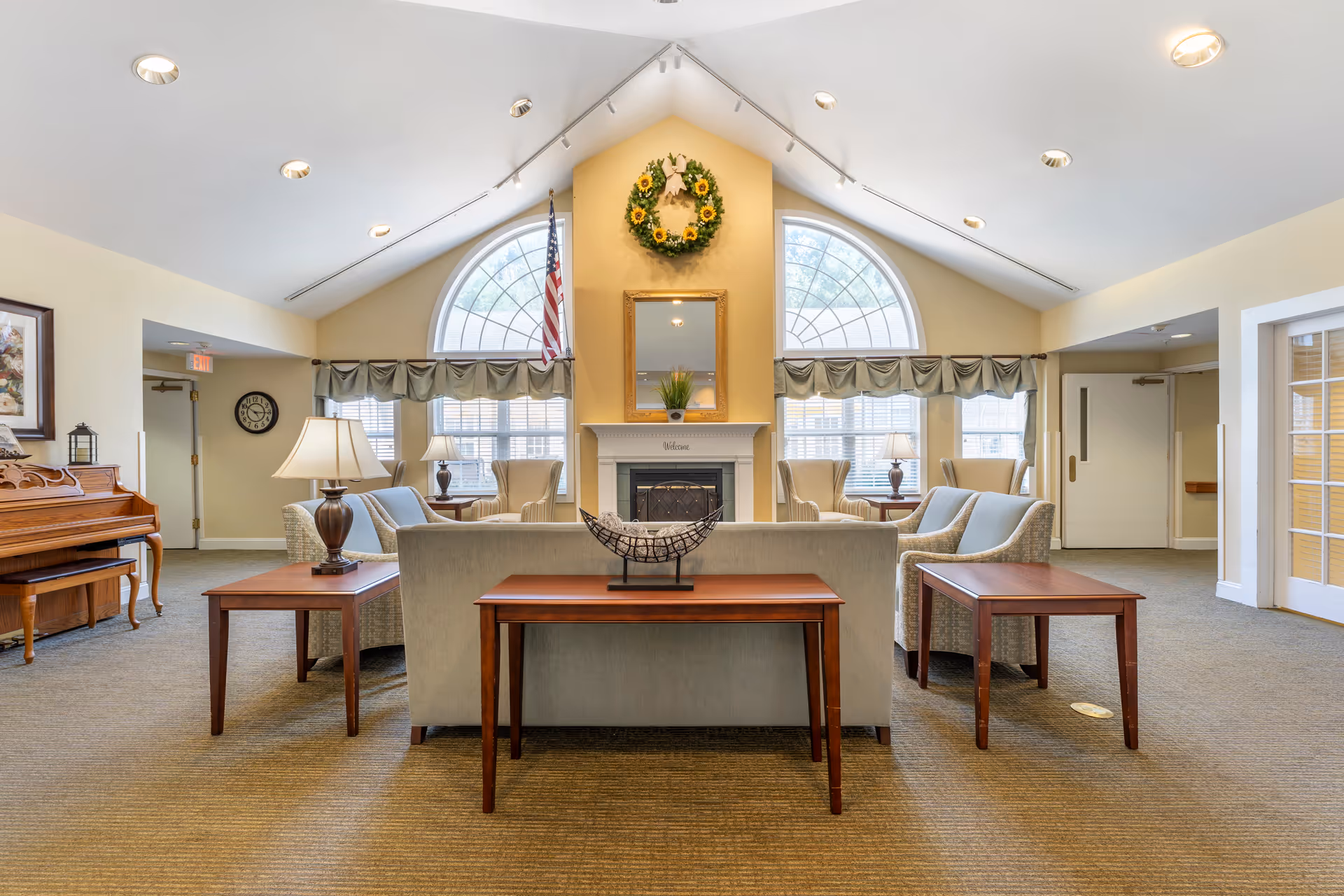 Common sitting area with sofas, armchairs, wooden side tables and a fireplace beneath arched windows and a decorative wreath.