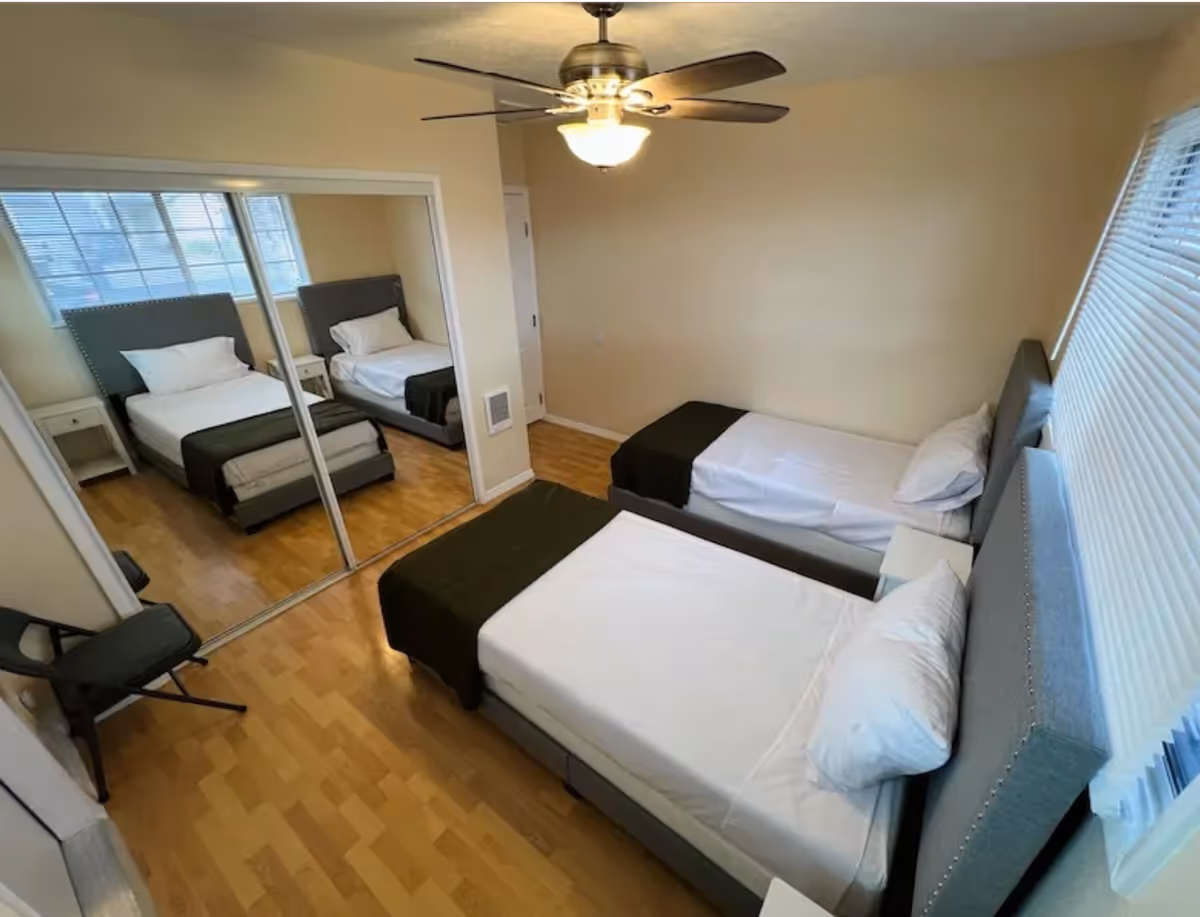 A sunlit bedroom with two twin beds, gray headboards, a ceiling fan, and a mirrored closet.