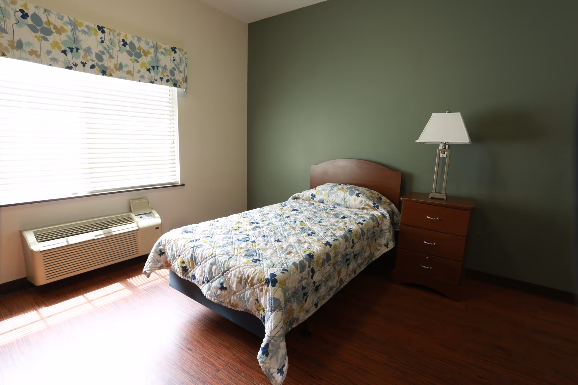 A simple bedroom with a single bed covered in a floral quilt, a wooden nightstand with a lamp, a window and an air conditioner against a green accent wall.