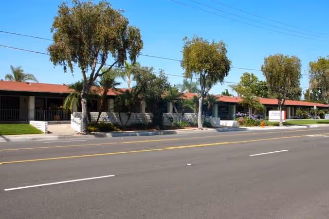 Street view of a single-story post-acute care building with a red roof, shaded walkways, trees and landscaping set back from a multi-lane road.