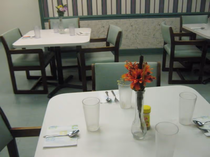A dining room with several tables and chairs arranged neatly. Each table has clear drinking glasses, utensils, napkins, and a small vase with flowers as a centerpiece. The room has a green and white color scheme with striped wallpaper and a green floor.
