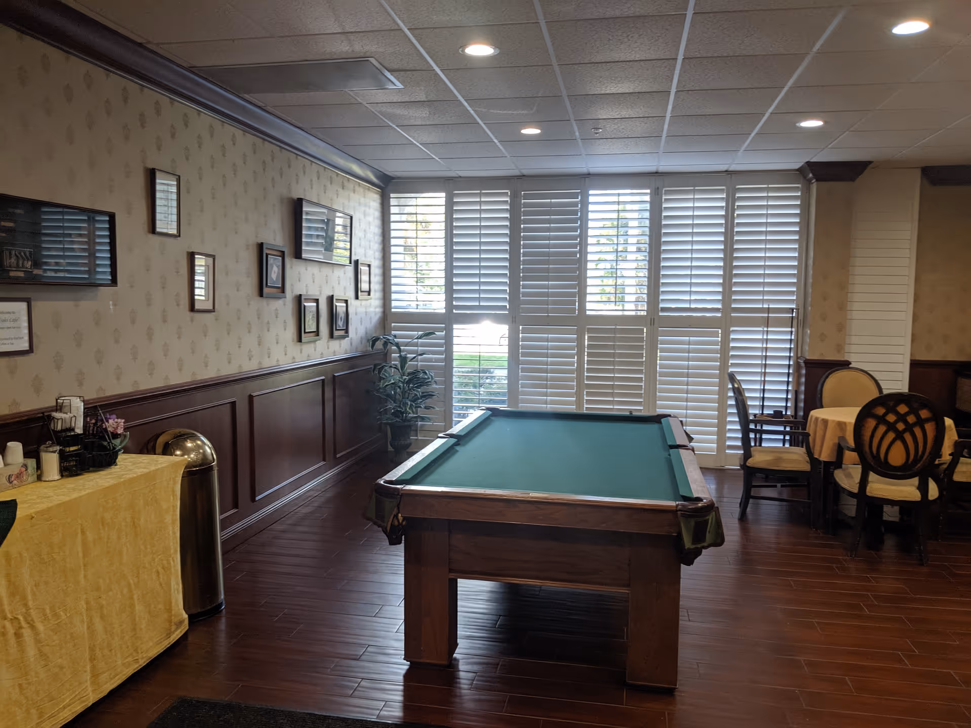 Indoor senior living recreation room featuring a pool table, seating, and large shuttered windows.