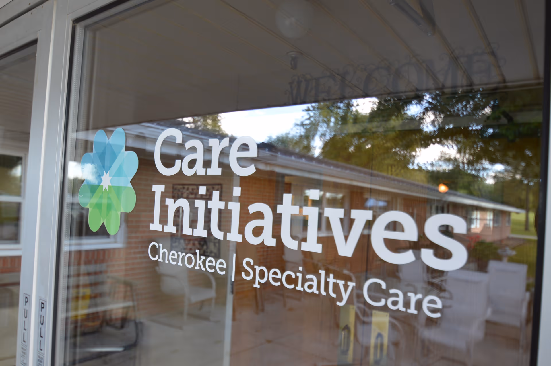 Glass door with the logo and text 'Care Initiatives Cherokee Specialty Care' printed on it, reflecting the outdoor area with trees and a brick building.
