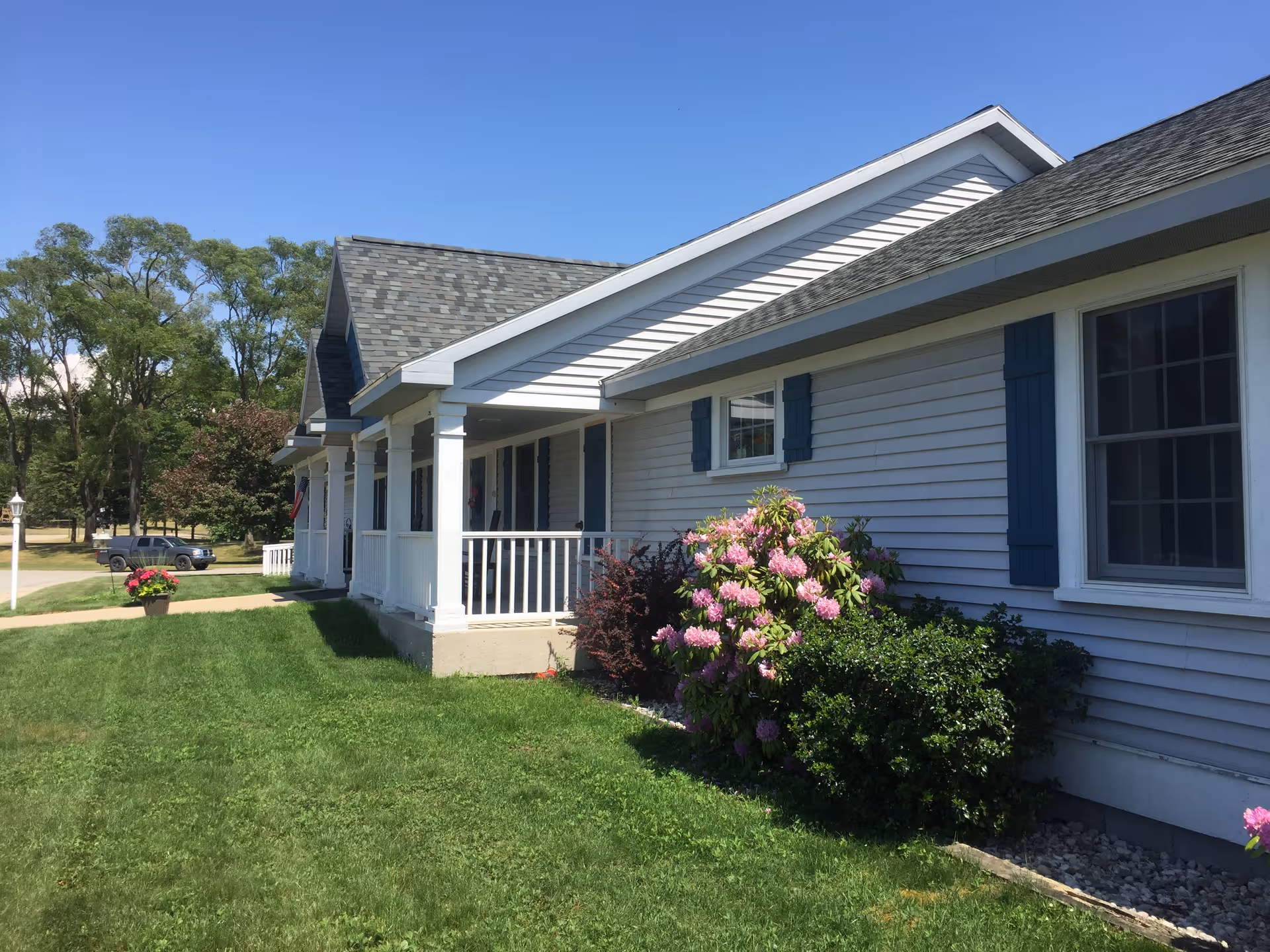 Exterior view of a single-story residential building with white siding and blue shutters, a covered porch with white railings, green lawn, flowering bushes, and trees under a clear blue sky.