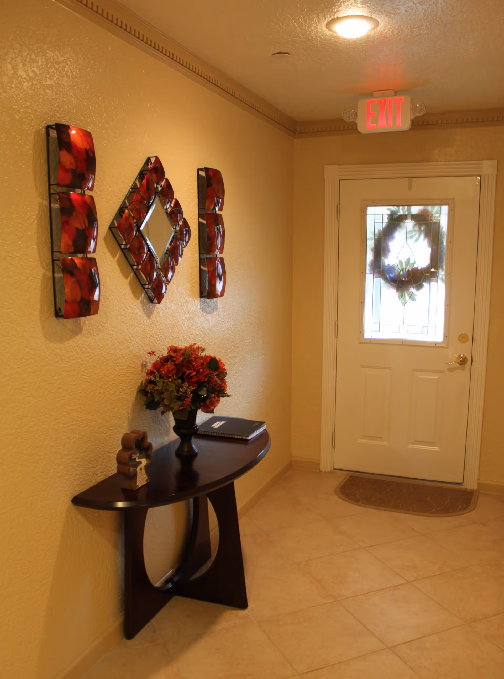 A hallway with beige walls and tiled floor featuring a dark wooden console table with a vase of red flowers and a book on it. Above the table is a decorative wall mirror with red accents. At the end of the hallway is a white door with a glass panel decorated with a wreath and an illuminated red exit sign above it.