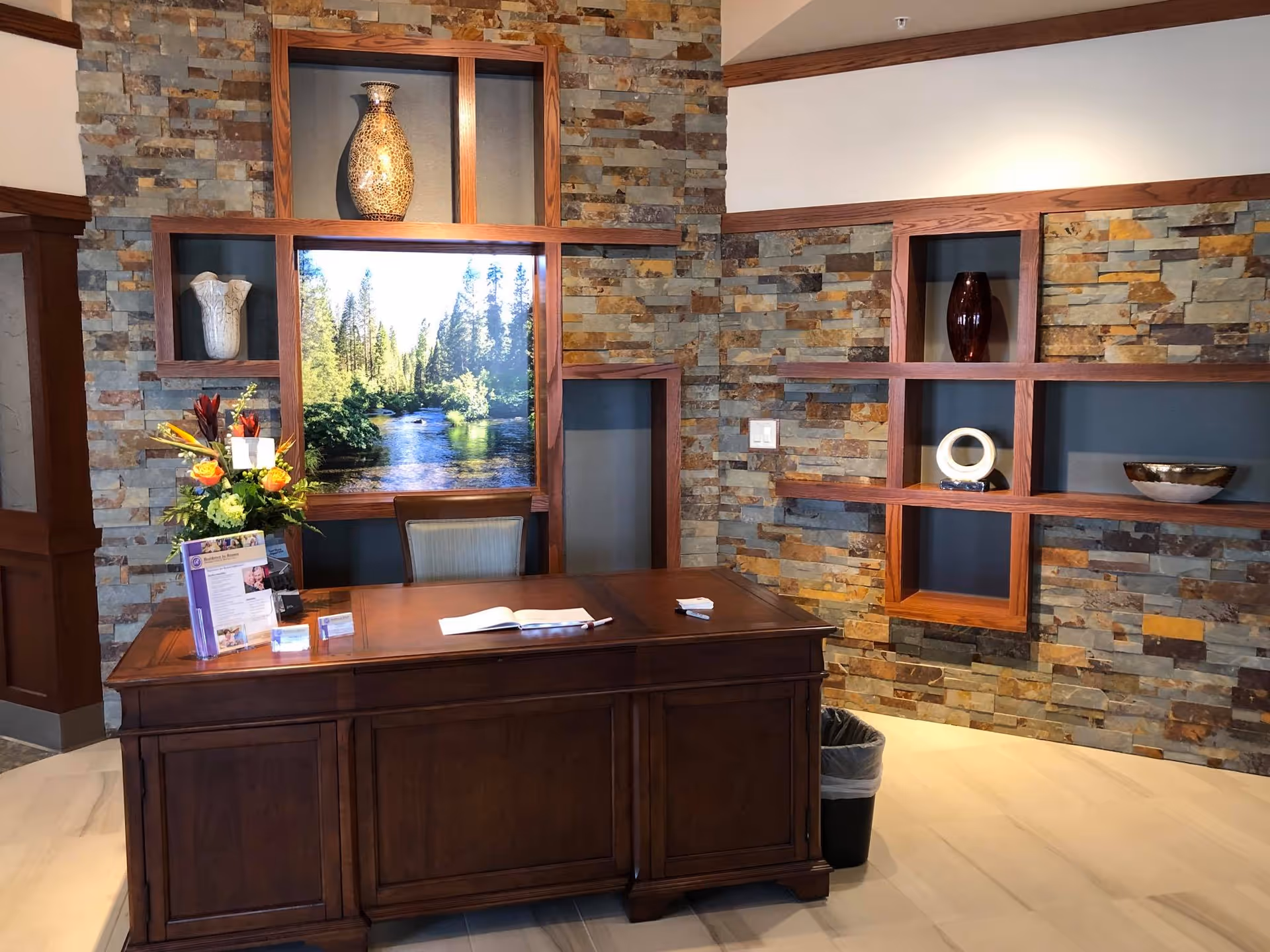 A reception desk area with a wooden desk in front of a stone accent wall featuring built-in wooden shelves displaying decorative vases and bowls. On the desk, there is a flower arrangement, informational brochures, and an open book. A framed picture of a river and trees is centered on the wall behind the desk.
