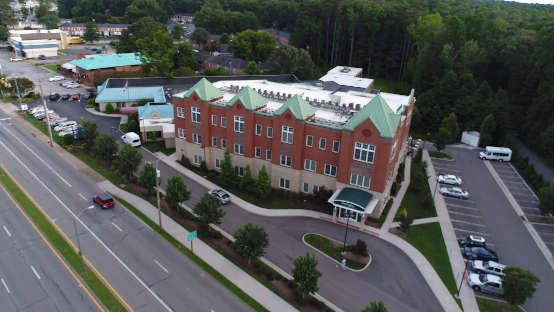 Aerial view of a multi-story brick building with green roofs, surrounded by trees and parking lots, located next to a multi-lane road with cars. The building has a driveway with a covered entrance and landscaped greenery around it.