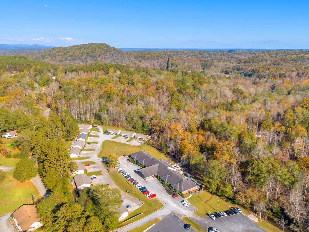 Aerial view of Overlook Toccoa facility surrounded by dense forest and rolling hills under a clear blue sky. The facility includes a main building with a parking lot and several smaller buildings or units nearby, all set in a rural, wooded landscape.