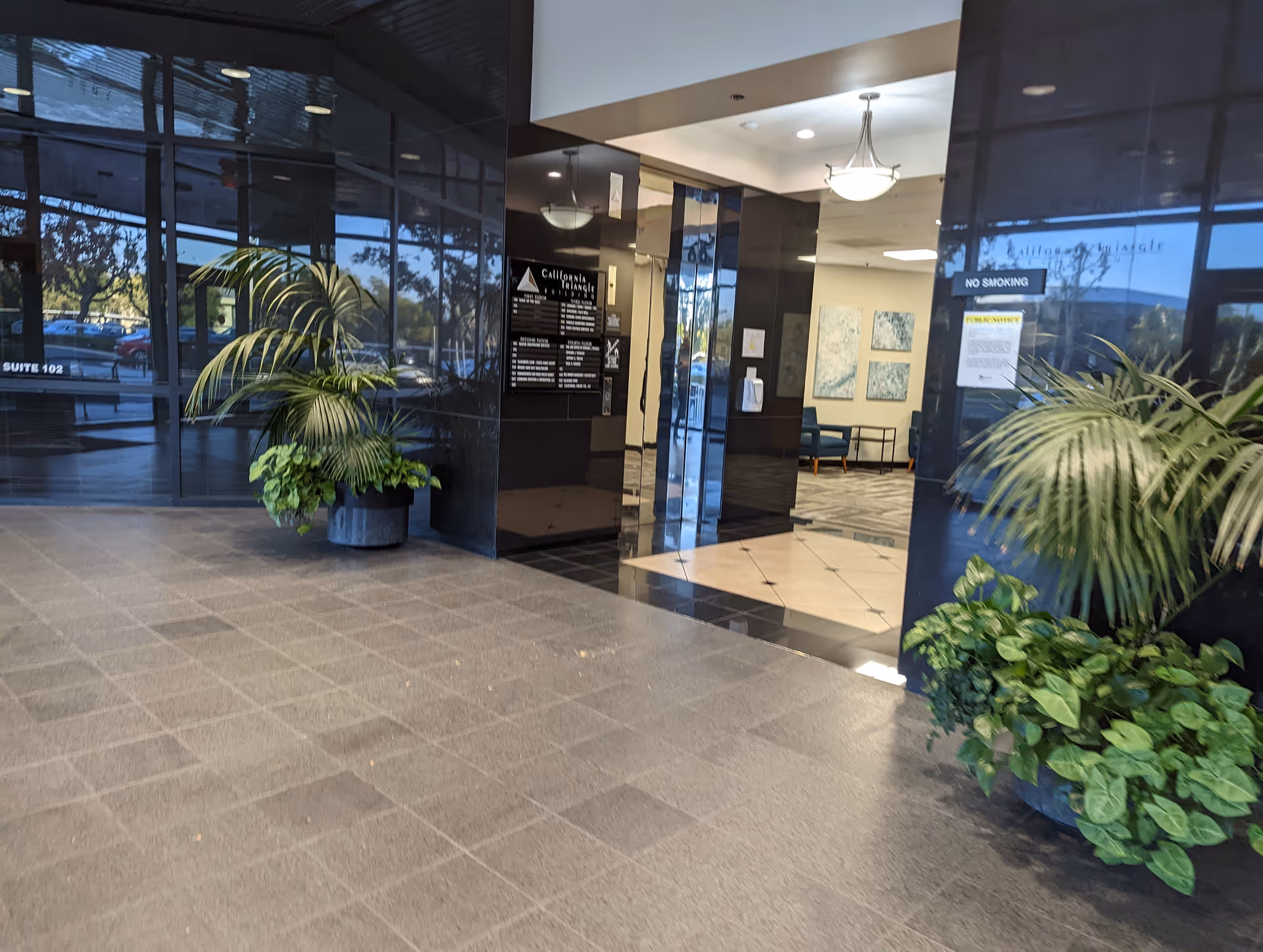 Modern building lobby with glass doors, potted plants, and checkered tile floor.
