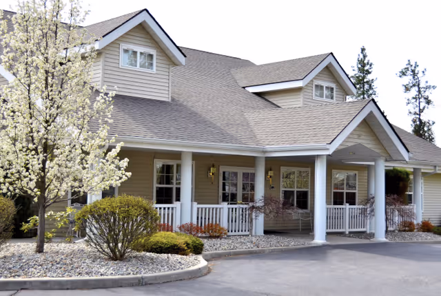 Front entrance of a light-colored senior living building with a covered porch, white columns, and landscaped trees and shrubs.