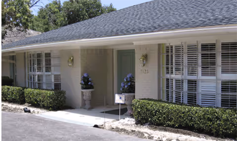 Single-story light-brick building exterior with front entrance, windows, hedges, and flowering planters.
