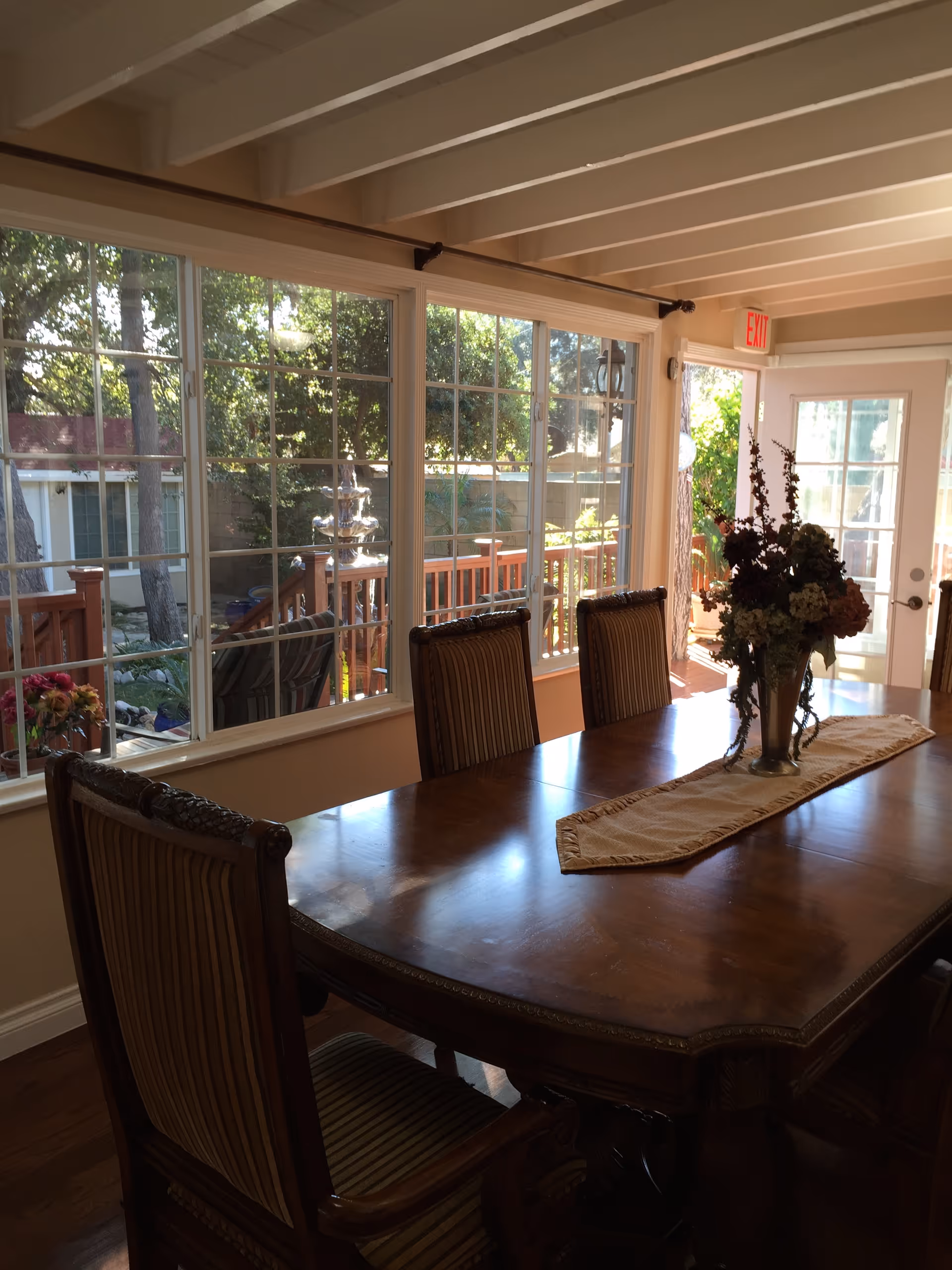 A dining room with a large wooden table and four upholstered chairs. The table has a decorative runner and a vase with flowers. Large windows on one side let in natural light and show an outdoor deck area with trees and plants. A door with glass panels leads outside, and an exit sign is visible above the door.