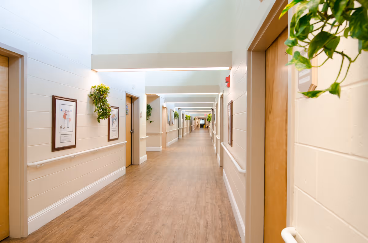 A long, well-lit hallway in a healthcare facility with wooden doors on both sides, framed pictures and green plants hanging on the walls, and wooden flooring.