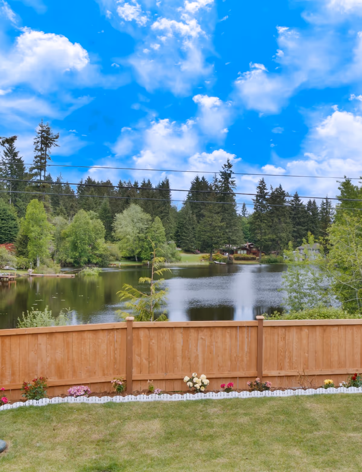 Lakeside backyard with a wooden fence, lawn, flowers, and a pond under blue sky.
