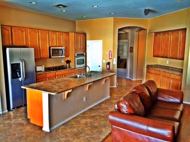 Open-plan kitchen with a center island, stainless steel refrigerator, wooden cabinets, and a brown leather sofa against the island.