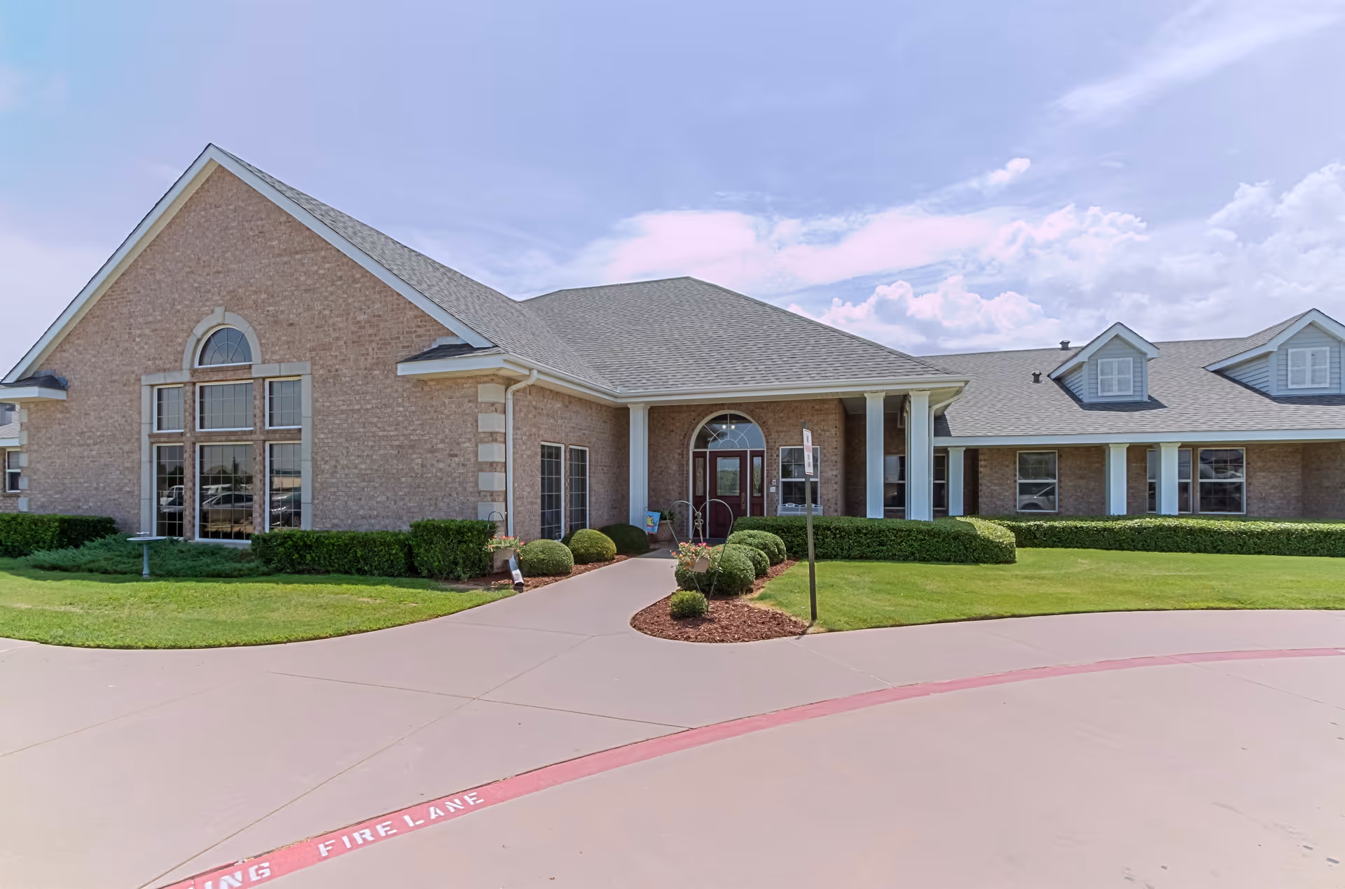Exterior front view of a single-story brick building with a gabled roof, large windows, and a covered entrance supported by white columns. The building is surrounded by neatly trimmed bushes and green grass, with a curved driveway marked with a red fire lane.