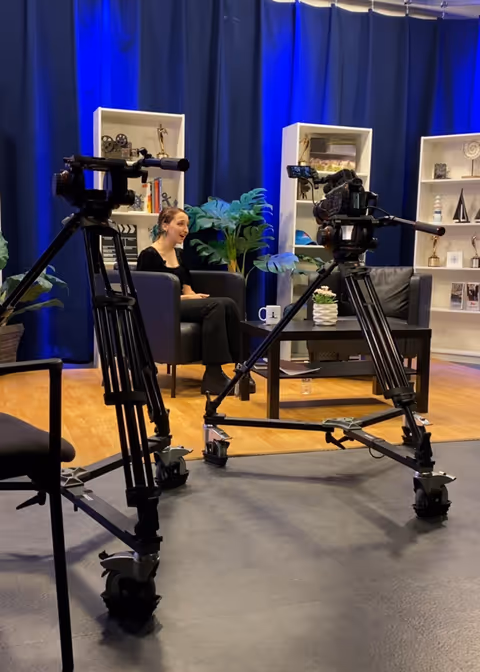 Studio interview setup with two cameras, a seated woman, blue curtains, and decorative shelves.