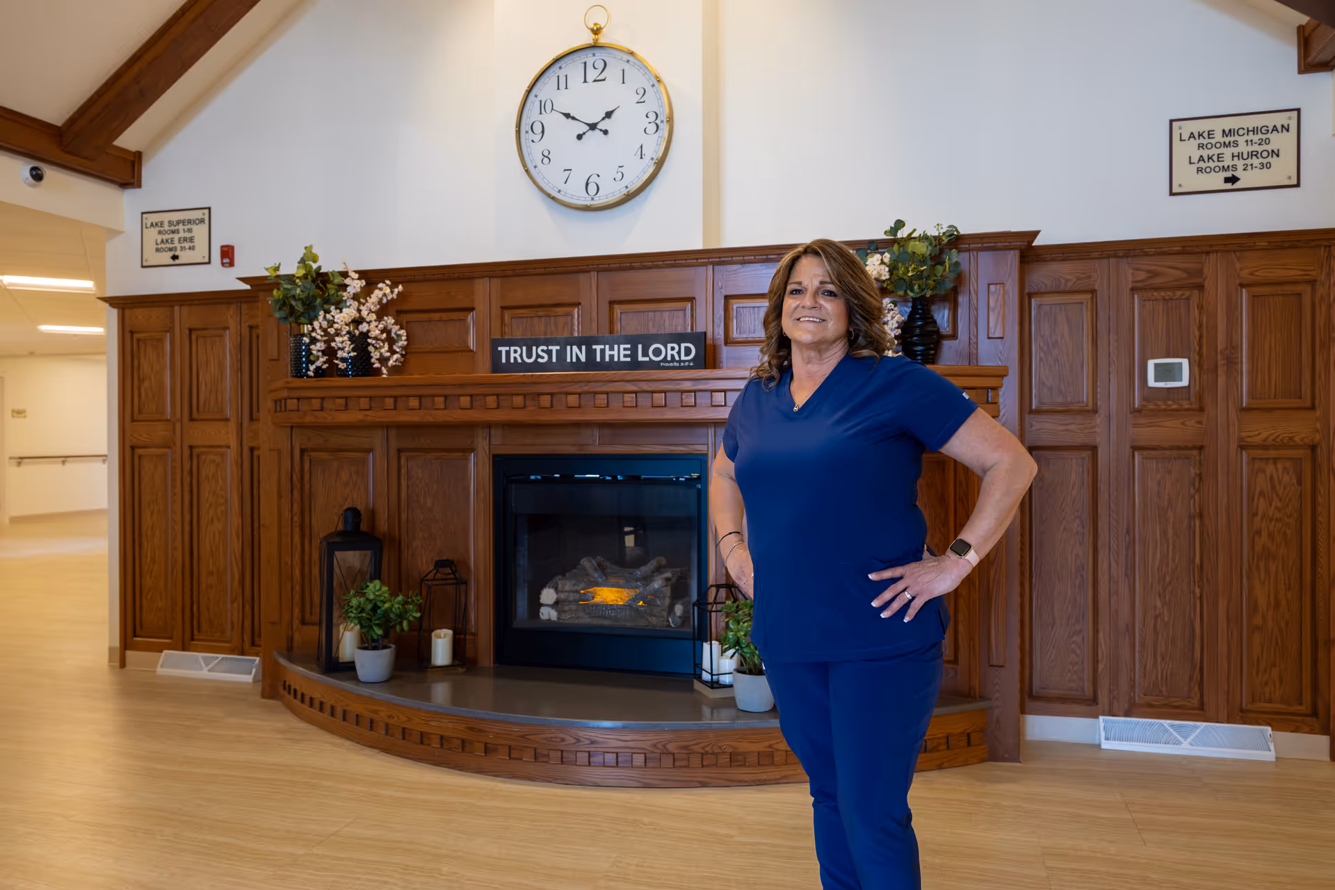 A woman in blue scrubs stands confidently with her hands on her hips in a room with wooden paneling and a fireplace. Above the fireplace is a clock showing the time as 10:10 and a sign that reads 'TRUST IN THE LORD'. There are decorative plants and candles on the mantle. Signs on the walls indicate directions to rooms named after lakes, such as Lake Superior, Lake Erie, Lake Michigan, and Lake Huron.