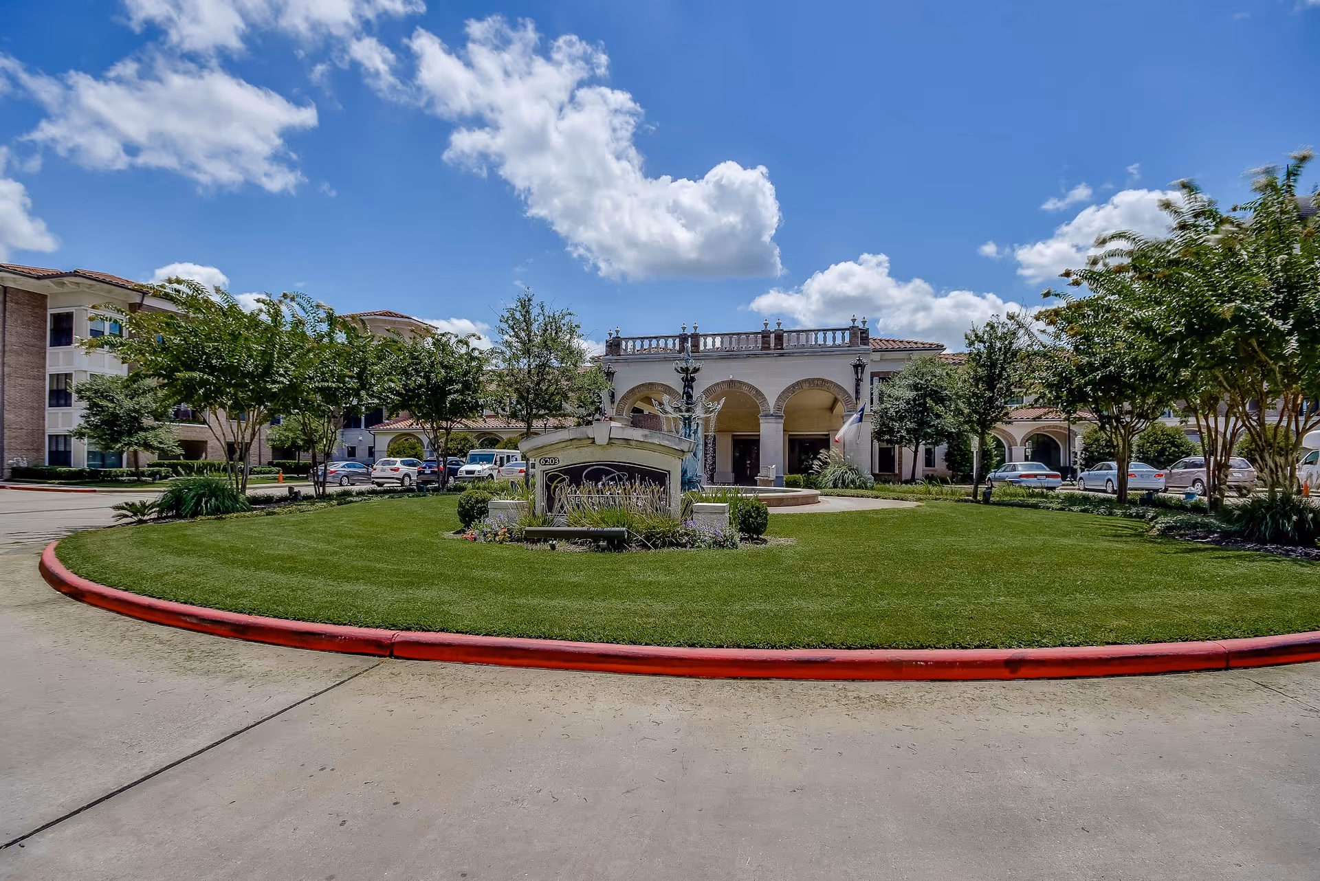 Front exterior view of Conservatory At Alden Bridge facility with a circular driveway, green lawn, trees, and a fountain in the center under a blue sky with scattered clouds.