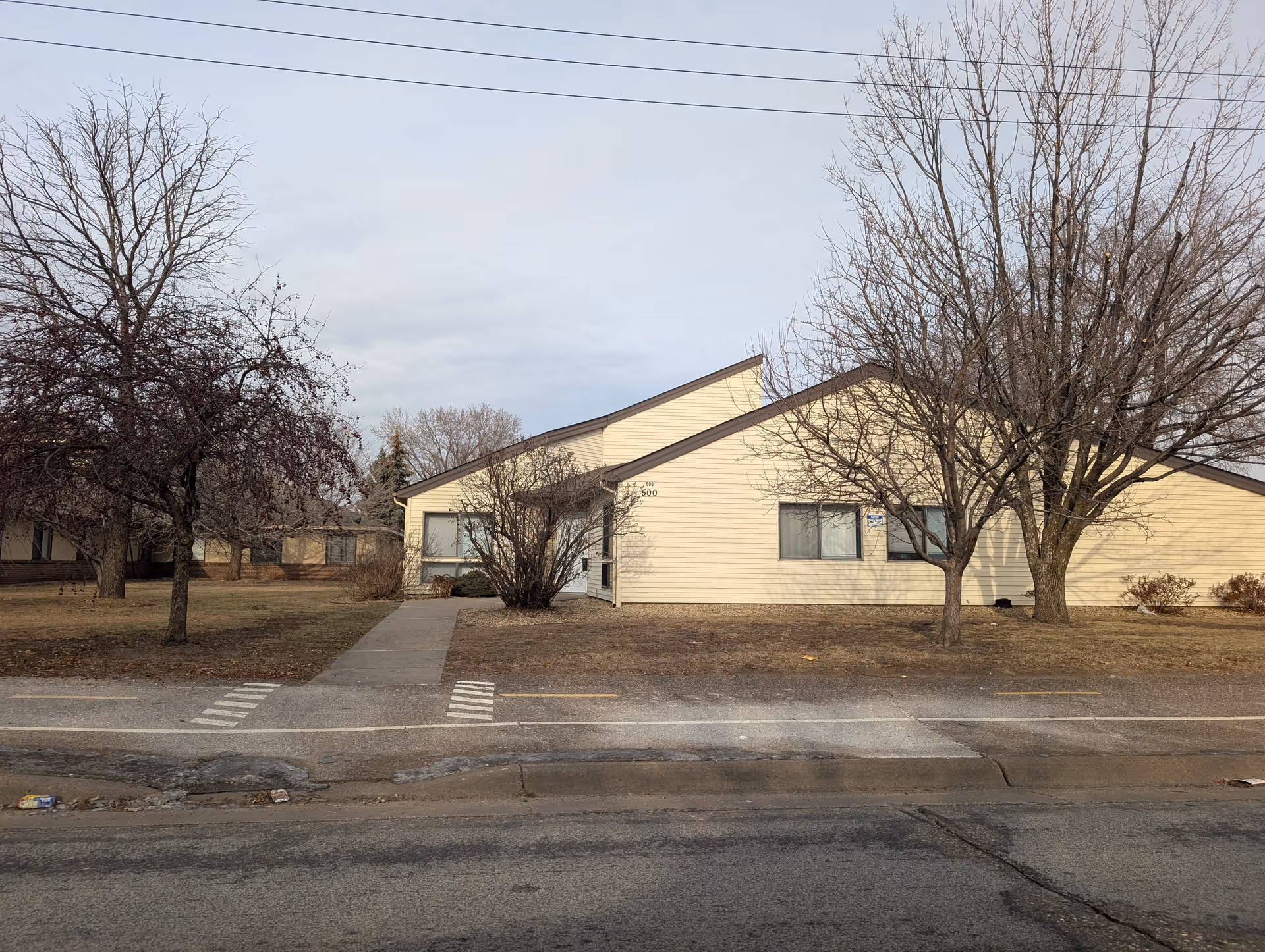 Front exterior of a single-story light-yellow building with bare trees, a sidewalk leading to the entrance, and a road in the foreground.