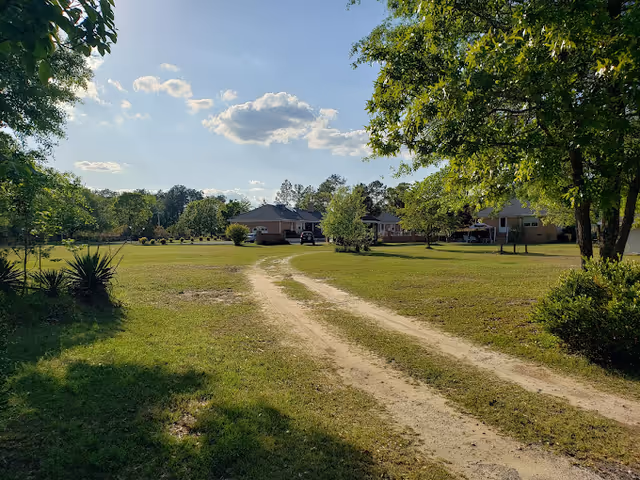 Dirt driveway winds through green lawn toward a single-story brick assisted living facility.