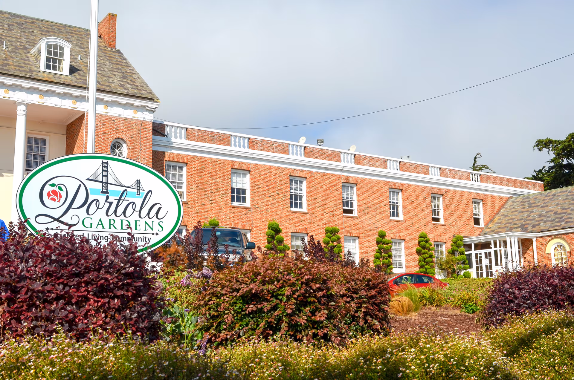 Exterior view of a brick building with multiple windows and a slate roof, surrounded by well-maintained bushes and plants. A large sign in front reads 'Portola Gardens Assisted Living Community' with an illustration of a bridge.