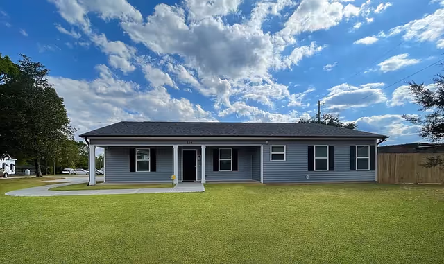 Single-story gray ranch-style house with a covered front porch, expansive green lawn, and a partly cloudy blue sky.