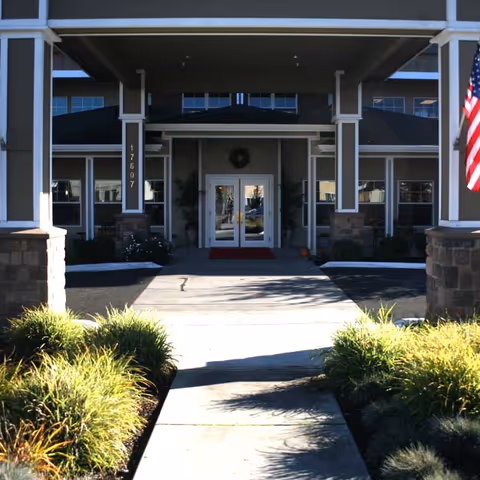 Covered main entrance with a walkway leading to double glass doors under a portico, flanked by columns and landscaping.