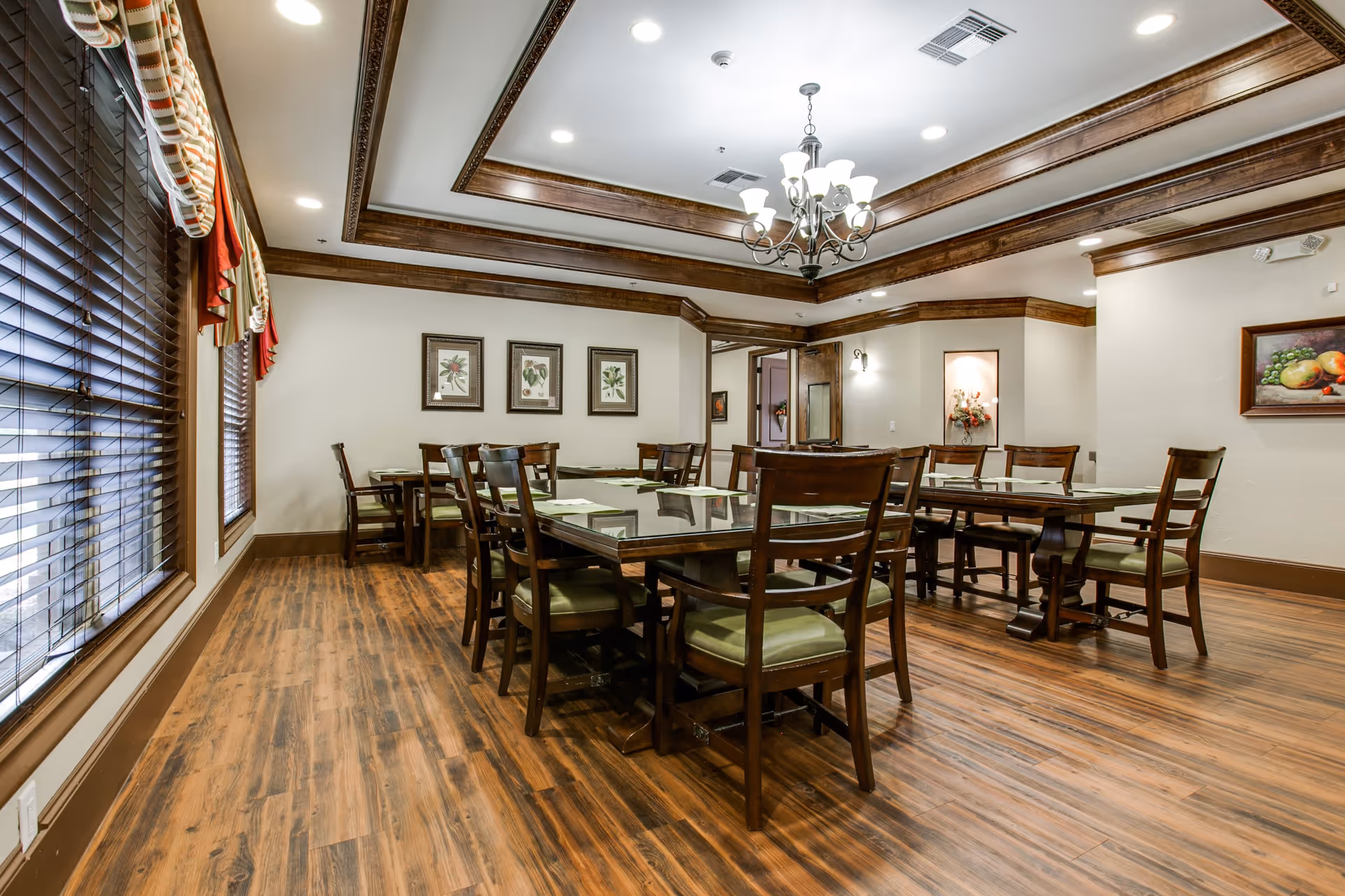 A well-lit dining room with wooden floors and multiple wooden tables and chairs arranged neatly. The room features large windows with blinds and valances, framed botanical artwork on the walls, and a decorative chandelier hanging from a coffered ceiling with wooden trim.